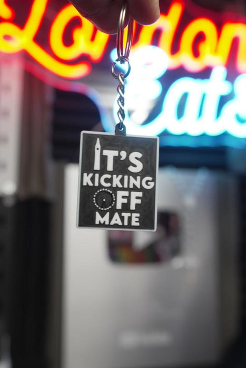 A hand holds a square metal keychain with a black and white graphic that reads 'IT'S KICKING OFF MATE' against a blurred neon sign background.