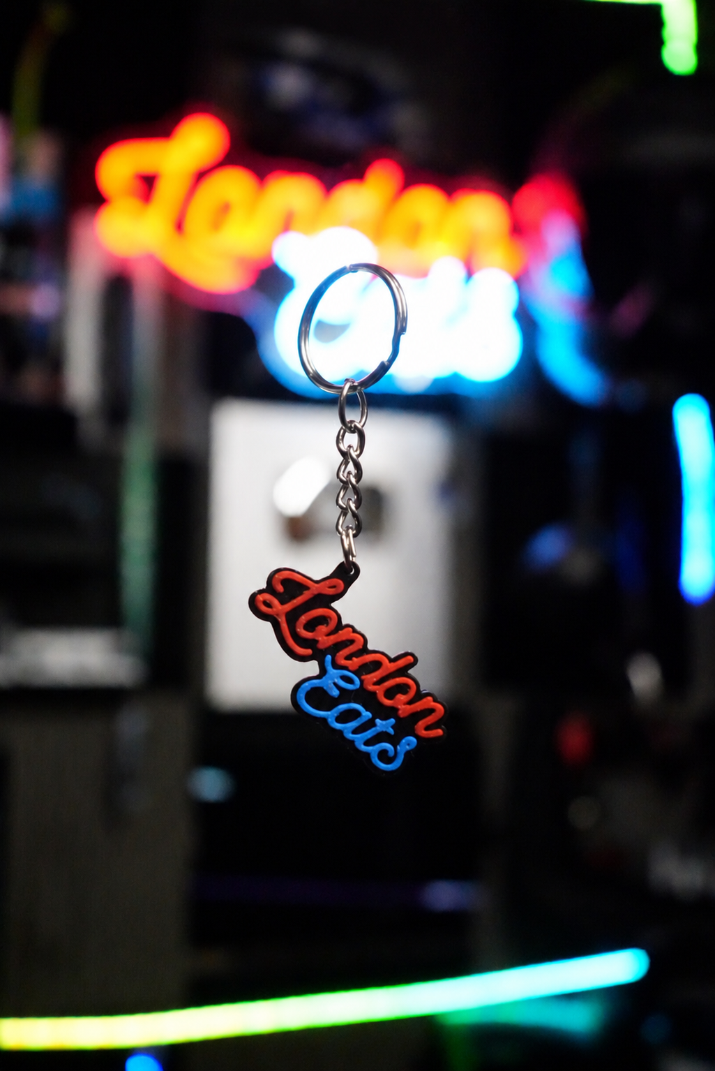 A small keychain with a silver split ring and chain, featuring a neon-style 'London Eats' sign in red and blue, photographed against a blurred neon-lit background.
